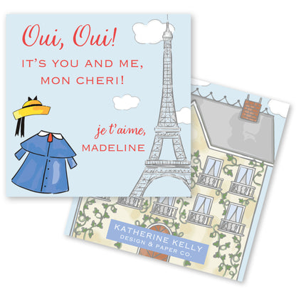 The image is a greeting card featuring a blue dress and a yellow hat, along with an illustration of the Eiffel Tower. The card has a French phrase "Ou, Ou! It's you and me, mon cheri!" and an illustration of a building with a chimney. The card also includes the text "KATHERINE KELLY DESIGN & PAPER CO."
