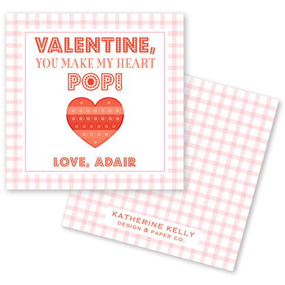 The image is a Valentine's Day card with a pink and white checkered background. The card features a heart-shaped pop-up toy and a message that reads "Valentine, You make my heart POP!" The card also includes the text "Love, Adair" and "KAT" at the bottom.
